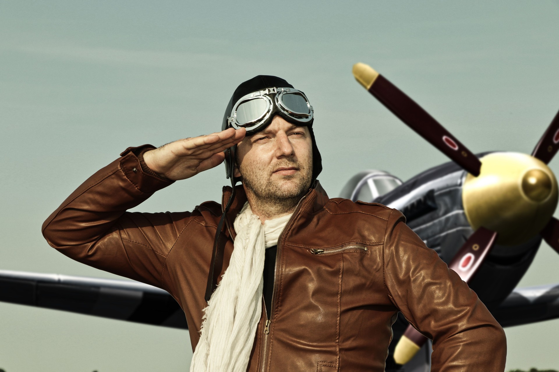 Portrait of a vintage pilot with leather cap, scarf and aviator glasses salutes in front of a historic airplane - Portrait of a man in historical pilot clothing